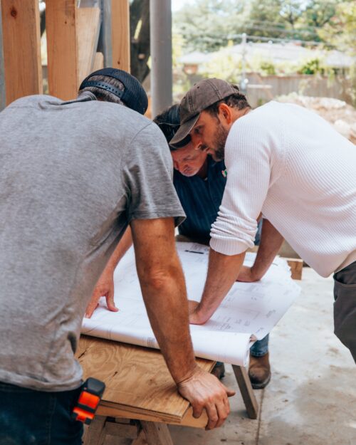 Two construction workers look at a blueprint on a bench. 